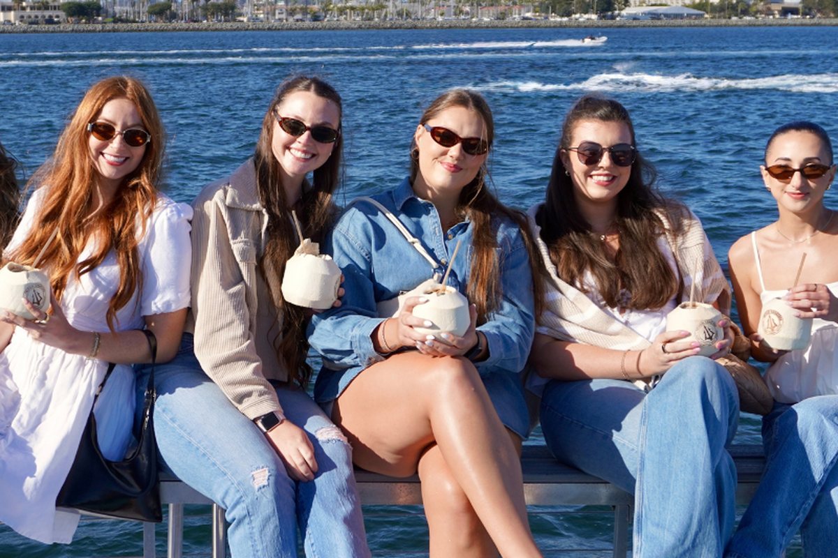 Five friends holding coconut drinks on Athena's rail seating, the bay and Coronado behind them
