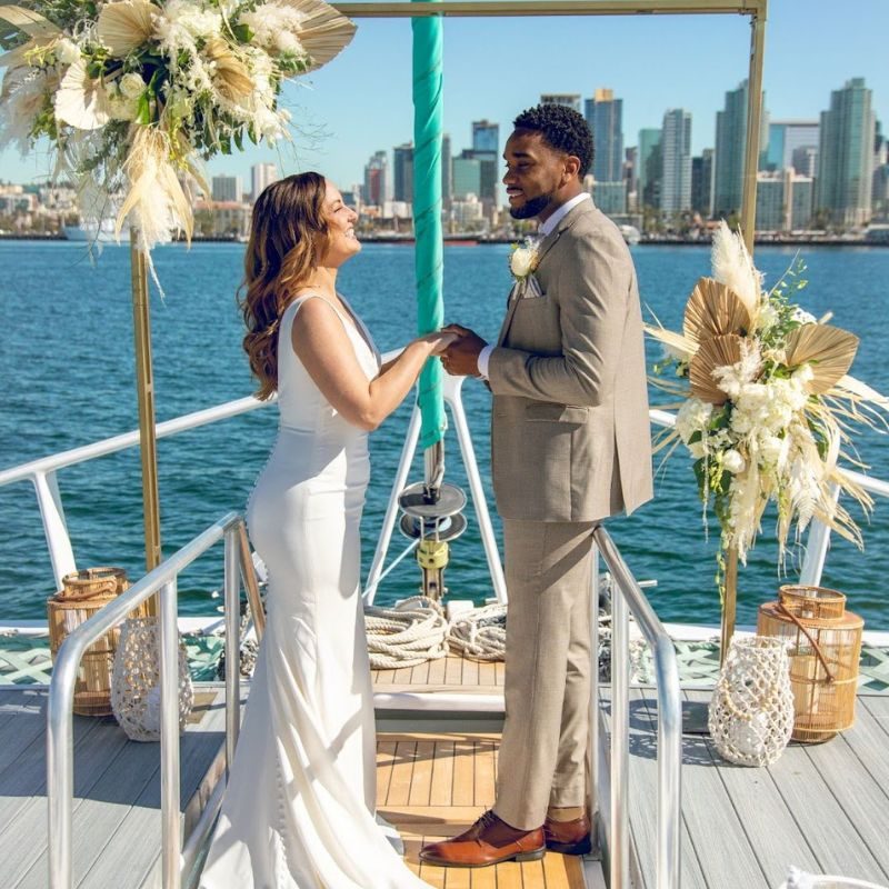 Couple exchanging vows under a pampas grass arch on the Triton’s deck with the San Diego skyline behind them