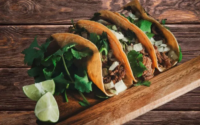 Three carne asada tacos lined up on a wooden board with a wedge of lime