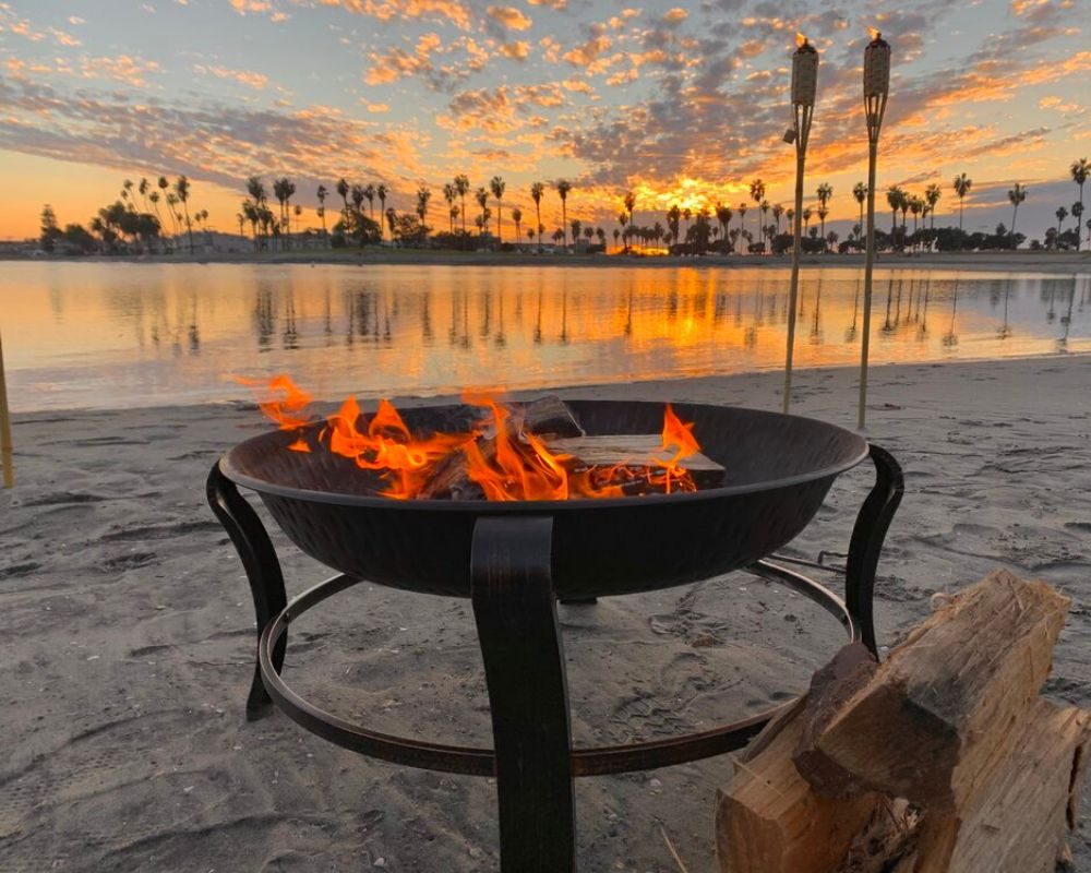 Beach fire pit burning at sunset with tiki torches and firewood beside it